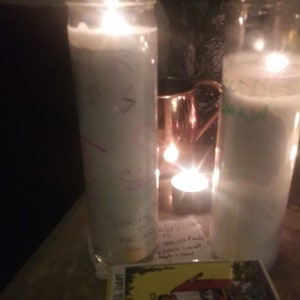 May include: Two lit white candles in glass jars, with handwritten notes on one. A tarot card deck with the "THE RIDER TAROT" label is in the foreground. A copper mug and a dark sculpture are in the background.