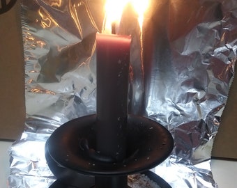 May include: A lit, dark red candle in a black metal candlestick. The candle is burning brightly, with flames reflecting off the aluminum foil behind it. The candlestick sits on a bed of white crystals and wood shavings.