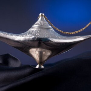 Könnte beinhalten: Eine silberfarbene Wunderlampe mit geschwungenem Ausguss und aufwendigen Details. Die Lampe hat einen dekorativen Deckel und eine goldfarbene Kette am Griff. Sie ist vor einem dunkelblauen Hintergrund platziert.