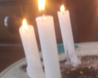 May include: Three white pillar candles with lit flames. The candles are on a white plate with a dark brown surface. The flames are bright yellow and orange, creating a warm, inviting ambiance. The candles are of varying heights.