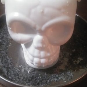 May include: A white skull-shaped candle rests in a silver metal dish containing dark grey granules. The candle features detailed facial characteristics, including eye sockets, a nose, and teeth. The background is a dark, wooden surface.