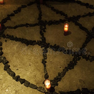 May include: A pentagram symbol made of dark stones on a concrete surface, with lit candles at each point. The candles emit a warm, orange glow, creating a mystical atmosphere. The arrangement suggests a ritualistic or spiritual theme.