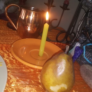 May include: A copper mug, a lit green candle in a yellow dish, and a pear rest on an orange tablecloth. The candle flame glows brightly. A black candelabra and other objects are in the background.