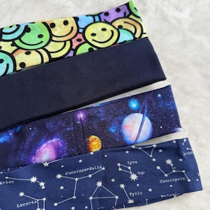 May include: Four headbands with various designs. The top one features a colourful smiley face pattern. Beneath it is a plain navy blue headband. The third headband has a galaxy design with planets and stars. The bottom headband is navy blue with white constellations.