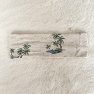 May include: A beige headband with a tropical palm tree design. The headband features a repeating pattern of palm trees, water, and small huts in shades of green, blue, and brown.