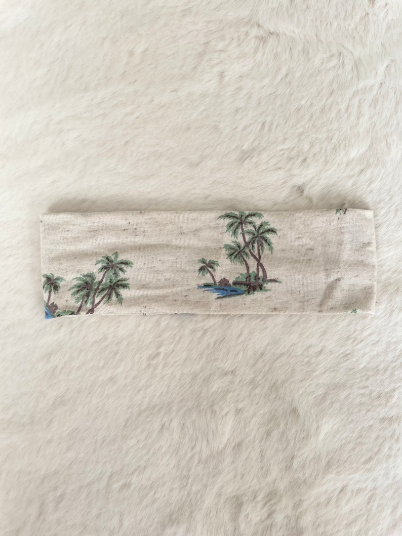 May include: A beige headband with a tropical palm tree design. The headband features a repeating pattern of palm trees, water, and small huts in shades of green, blue, and brown.