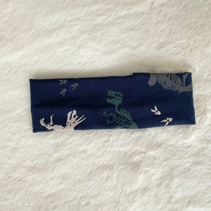 May include: A navy blue headband featuring a dinosaur skeleton and footprint pattern in gray and green. The fabric is soft and stretchy, suitable for various head sizes. The headband is a stylish accessory.