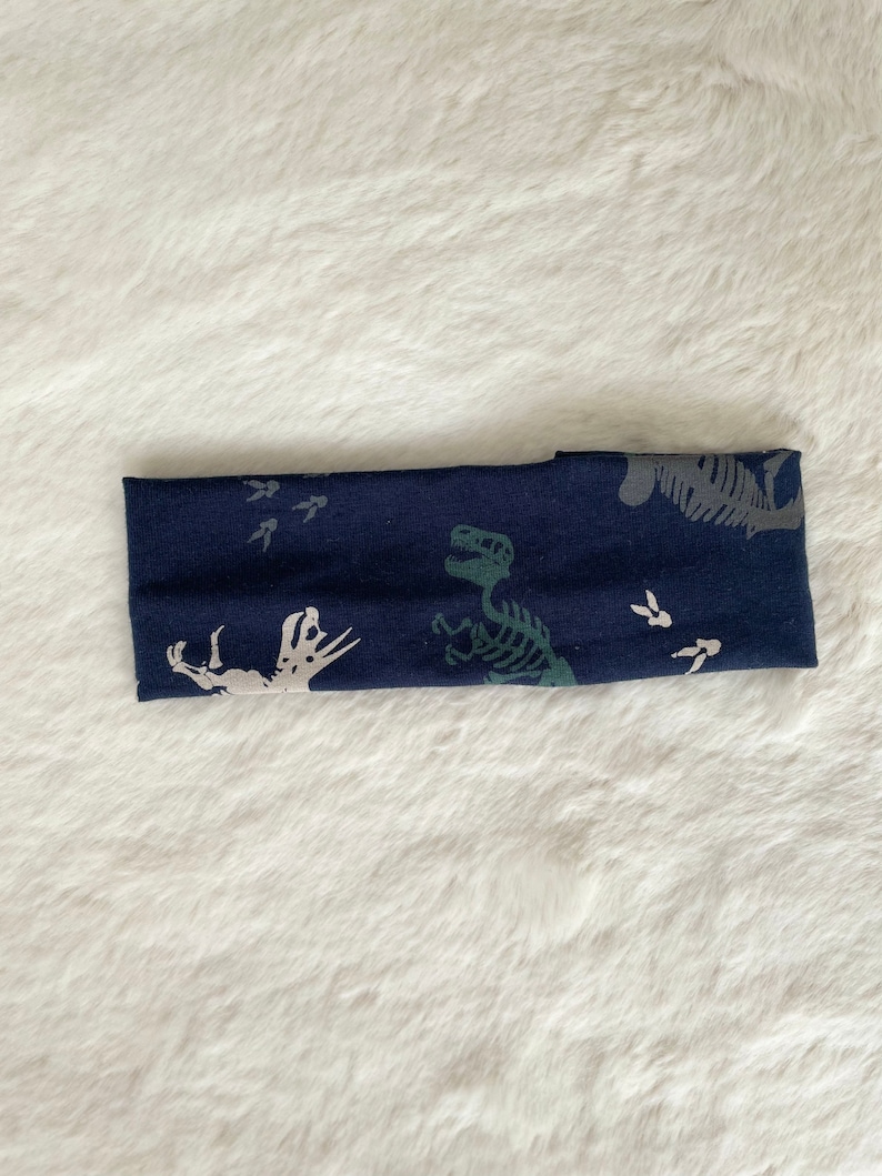 May include: A navy blue headband featuring a dinosaur skeleton and footprint pattern in gray and green. The fabric is soft and stretchy, suitable for various head sizes. The headband is a stylish accessory.
