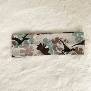 May include: A beige headband with a repeating pattern of brown dinosaurs and teal and brown leaf shapes. The headband is made of a soft, stretchy fabric and is designed to fit comfortably around the head.