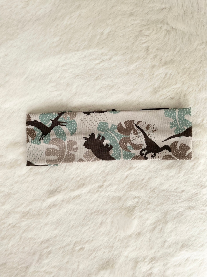 May include: A beige headband with a repeating pattern of brown dinosaurs and teal and brown leaf shapes. The headband is made of a soft, stretchy fabric and is designed to fit comfortably around the head.