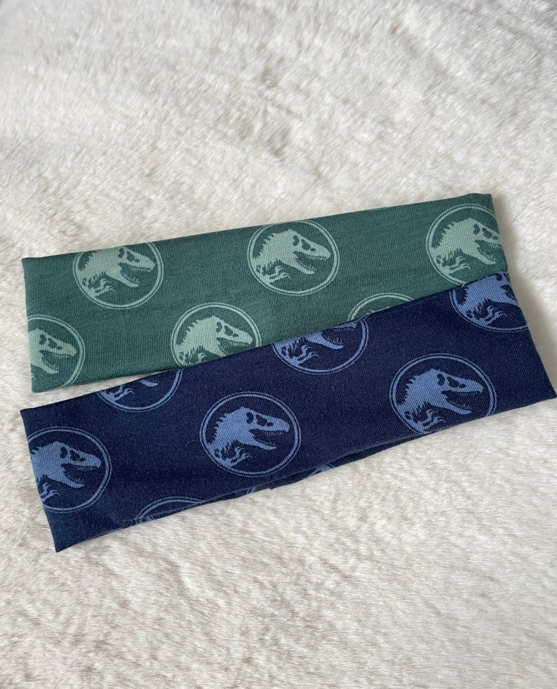 May include: Two headbands with a dinosaur pattern. One headband is green with a light blue dinosaur print. The other headband is dark blue with a light blue dinosaur print.