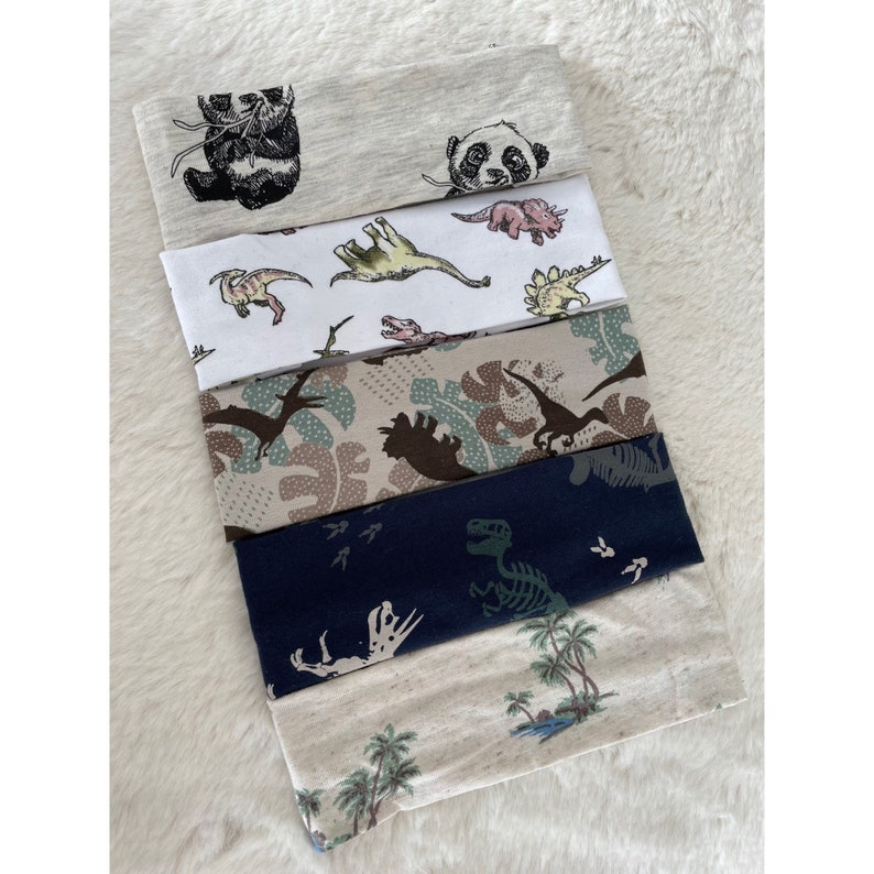May include: A stack of fabric headbands with various patterns. Designs include pandas, dinosaurs, and tropical scenes. The headbands are in shades of gray, white, beige, and navy blue.