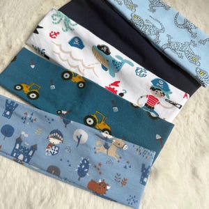Pode incluir: Uma coleção de bandanas coloridas com vários padrões. Os designs incluem lagartos azuis, um azul marinho liso, um tema pirata com uma baleia, tratores e um tema medieval com um castelo e um cavaleiro. As bandanas estão sobre uma superfície branca e fofa.