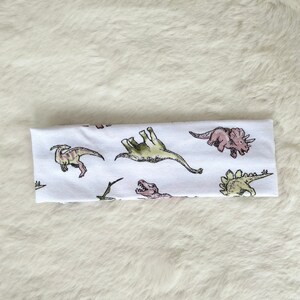 May include: White headband featuring a repeating pattern of various dinosaurs in shades of green, pink, and brown. The dinosaurs are illustrated in a whimsical style, creating a playful and fun design. The headband is made of a soft, likely stretchy material.
