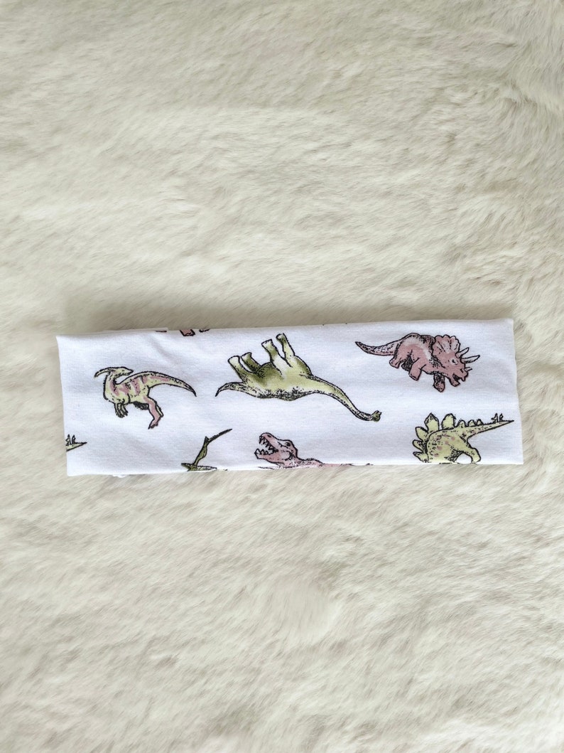 May include: White headband featuring a repeating pattern of various dinosaurs in shades of green, pink, and brown. The dinosaurs are illustrated in a whimsical style, creating a playful and fun design. The headband is made of a soft, likely stretchy material.