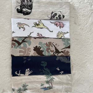 May include: A stack of six fabric headbands with various patterns. Designs include pandas, dinosaurs, camouflage, dinosaur skeletons, and tropical scenes with palm trees. The headbands are in shades of gray, white, blue, and beige.