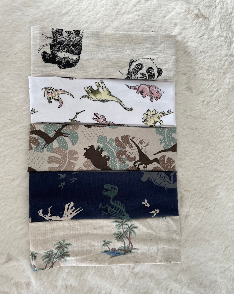 May include: A stack of six fabric headbands with various patterns. Designs include pandas, dinosaurs, camouflage, dinosaur skeletons, and tropical scenes with palm trees. The headbands are in shades of gray, white, blue, and beige.