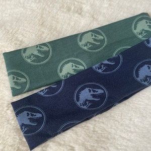 May include: Two fabric headbands with a dinosaur pattern. One headband is dark green with light green dinosaurs, and the other is dark blue with light blue dinosaurs.
