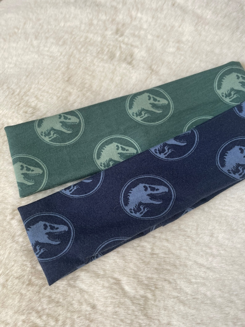 May include: Two fabric headbands with a dinosaur pattern. One headband is dark green with light green dinosaurs, and the other is dark blue with light blue dinosaurs.