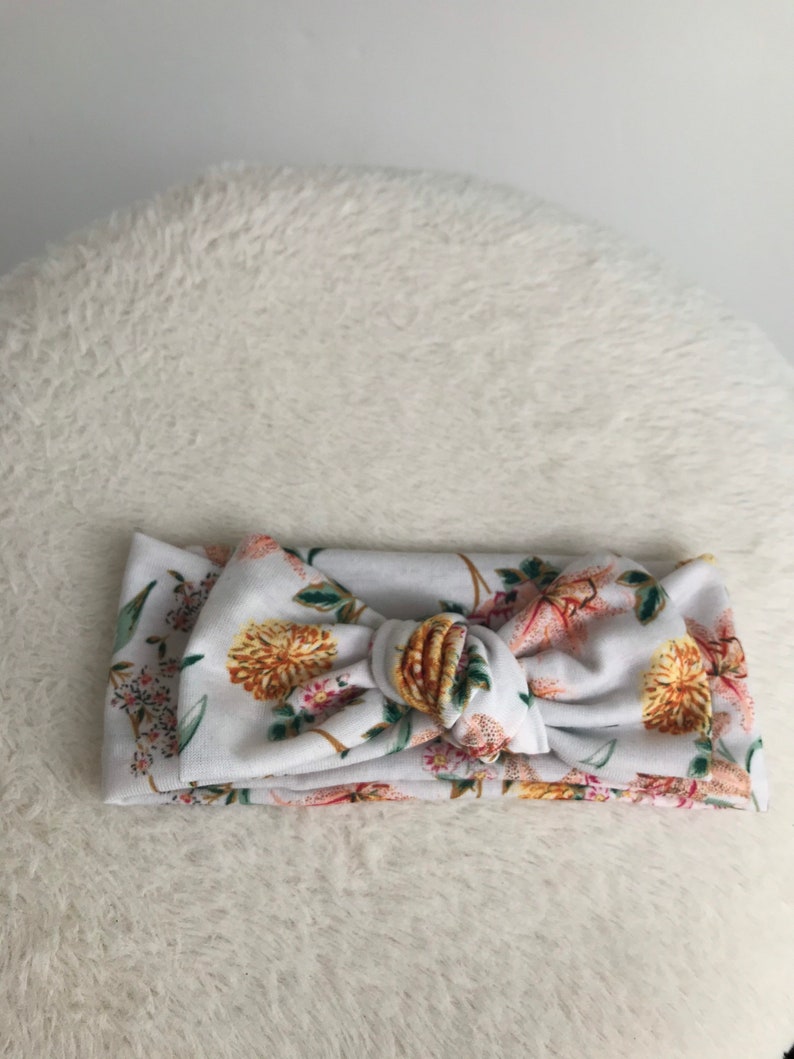 Pode incluir: Uma faixa de cabe&ccedil;a branca com estampa floral em tons de rosa, amarelo e verde. A faixa de cabe&ccedil;a &eacute; amarrada em um n&oacute; na parte superior.
