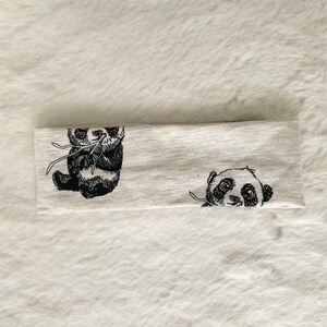 May include: A light gray headband featuring two black and white panda bear illustrations. One panda is sitting, and the other is peeking out. The headband is made of a soft, stretchy material.