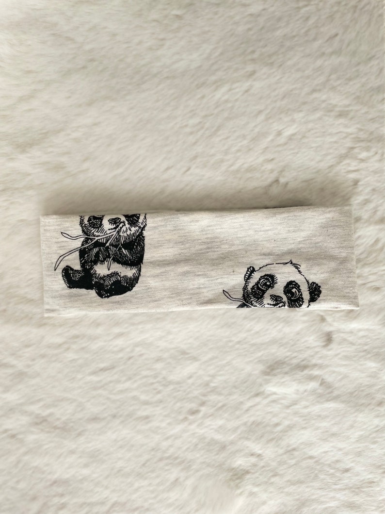May include: A light gray headband featuring two black and white panda bear illustrations. One panda is sitting, and the other is peeking out. The headband is made of a soft, stretchy material.