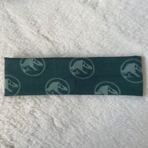 May include: A dark green headband with a repeating pattern of a white dinosaur silhouette inside a circle.