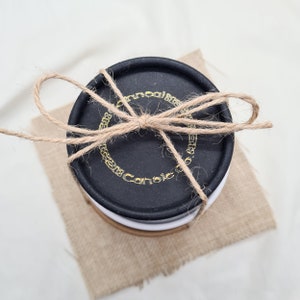 May include: A black round box with gold lettering that reads "Cannoli" and a brown twine bow. The box is sitting on a brown burlap surface.