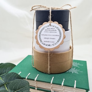 May include: Three stacked candle jars with brown paper lids and a white label with the text "Coinnear Candle Co." and a list of scents: "Earl Grey Tea & Cucumber, Midnight Berries, Rhubarb & Rose". The jars are tied together with twine.