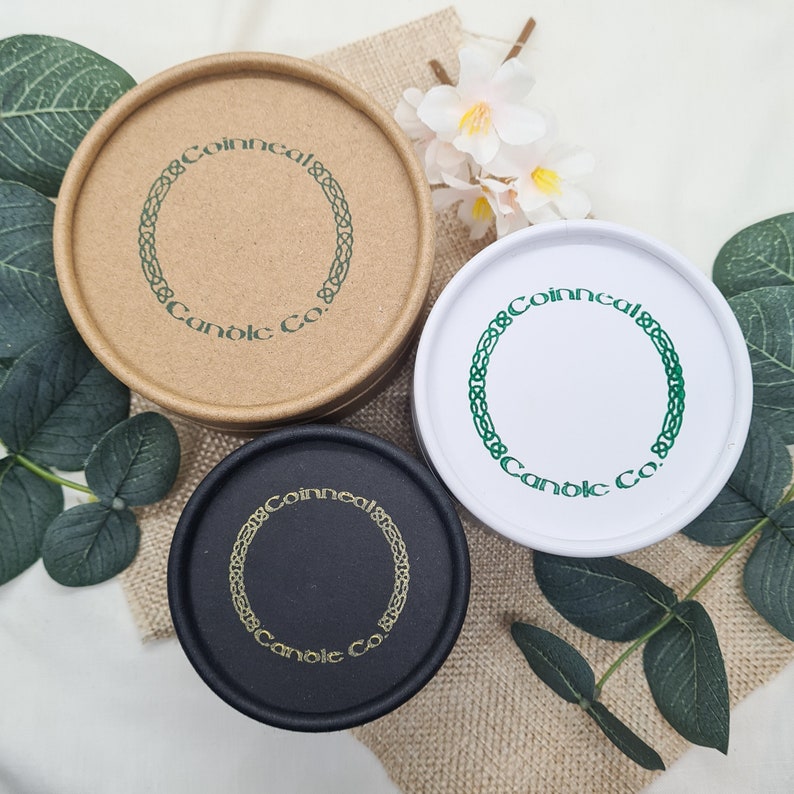 May include: Three round candle containers with lids. The containers are made of cardboard and are in brown, white, and black. Each container has a green Celtic knot design with the text "Coinneal & Candle Co."