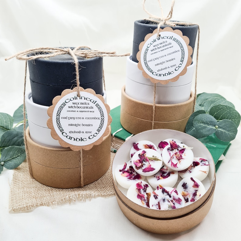 May include: A set of three brown cardboard boxes with black and white labels. The labels have text that reads "wax melts with botanicals" and "Cool grey tea & cucumber, midnight berries, shubarb & rose". The boxes are stacked on top of each other. A smaller brown cardboard box with a white lid contains wax melts with dried rose petals.