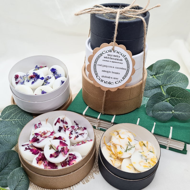 May include: Three round containers of wax melts with botanicals. The containers are stacked on top of each other. The top container is black, the middle container is white, and the bottom container is brown. The wax melts are in different colors and have different botanicals in them. The wax melts are labeled with the text "Coincal & Candle Co.", "wax melts with botanicals", "coconut & apricot wax", "cool grey tea & cucumber", "midnight berries", "rhubarb & anise".