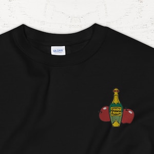 Peut inclure: Sweat-shirt noir avec une bouteille verte et dorée et deux pommes rouges brodées sur la poitrine gauche.