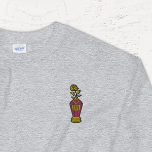 Könnte beinhalten: Ein graues Sweatshirt mit einem kleinen gestickten Design auf der linken Brust. Das Design zeigt eine rote Vase mit einer gelben Rose und goldenen Akzenten.
