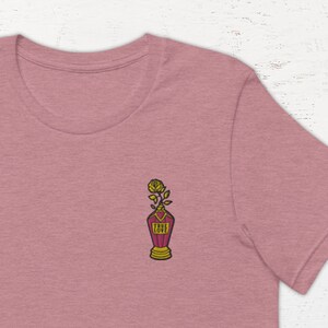 Könnte beinhalten: Ein heidefarbenes T-Shirt mit einem kleinen gestickten Design einer Rose in einer roten Vase mit dem Wort "LOVE" darauf.