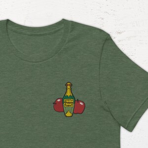 Peut inclure: Un t-shirt vert avec un petit motif brodé représentant une bouteille de cidre de pomme et deux pommes rouges.