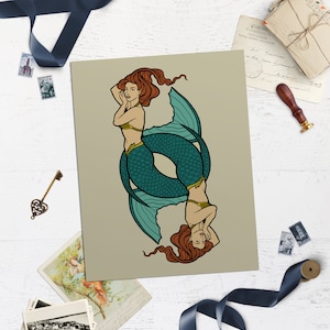 May include: A print featuring two symmetrical mermaids, one upright and one inverted. The mermaids have red hair, teal fishtails, and gold accents. The background is a neutral beige, and the artwork is surrounded by decorative elements.