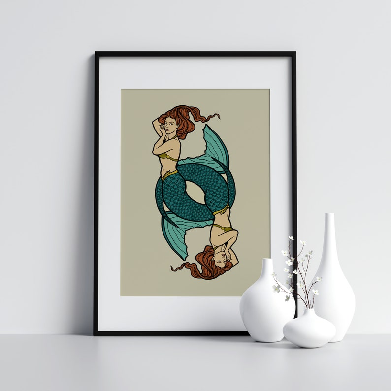 May include: A framed art print featuring a symmetrical illustration of two mermaids. The mermaids have long auburn hair, teal fishtails, and are wearing gold tops. The background is a muted beige color. The print is displayed with white vases.