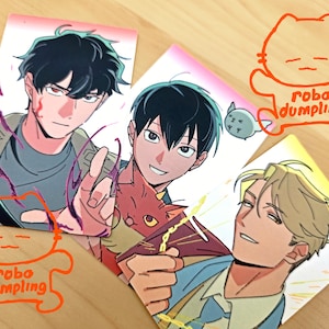 May include: Three colorful anime-style character art prints. The prints feature male characters with various expressions and poses. The prints are decorated with an orange cat-shaped logo that says "robo dumpling".