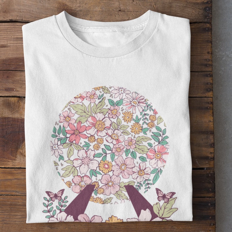 May include: White t-shirt with a floral print of a sphere. The sphere is surrounded by green leaves and two butterflies.