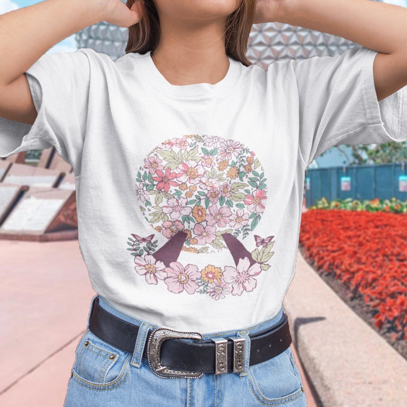 May include: White t-shirt with a floral print of a sphere, two butterflies, and a floral border. The sphere is reminiscent of a popular theme park attraction.