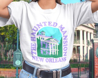 May include: White t-shirt with a purple and green graphic of the Haunted Mansion in New Orleans. The text "The Haunted Mansion New Orleans" is printed on the shirt.