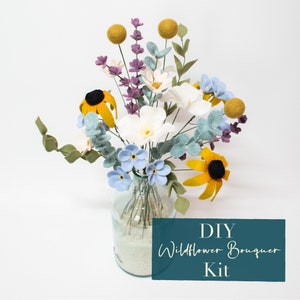 May include: DIY Wildflower Bouquet Kit featuring a felt flower arrangement in a glass vase with sand. The bouquet includes white, yellow, blue, and purple felt flowers.