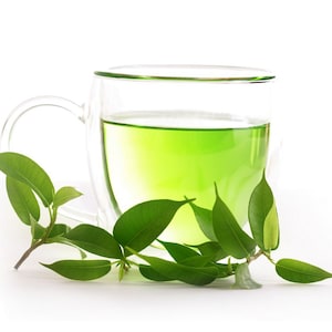Pode incluir: Uma caneca de vidro transparente cheia de l&iacute;quido verde, possivelmente ch&aacute;, &eacute; mostrada. Folhas e caules verdes frescos s&atilde;o dispostos ao redor da caneca. A caneca tem uma al&ccedil;a e um design de parede dupla. O fundo &eacute; branco.