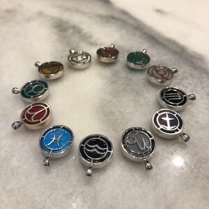 Op de afbeelding: Twaalf zilveren bedels met verschillende sterrenbeelden en kleuren. Elke bedel heeft een zilveren lus om aan een ketting of armband te bevestigen.