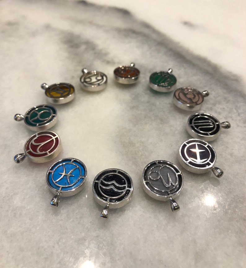 Op de afbeelding: Twaalf zilveren bedels met verschillende sterrenbeelden en kleuren. Elke bedel heeft een zilveren lus om aan een ketting of armband te bevestigen.