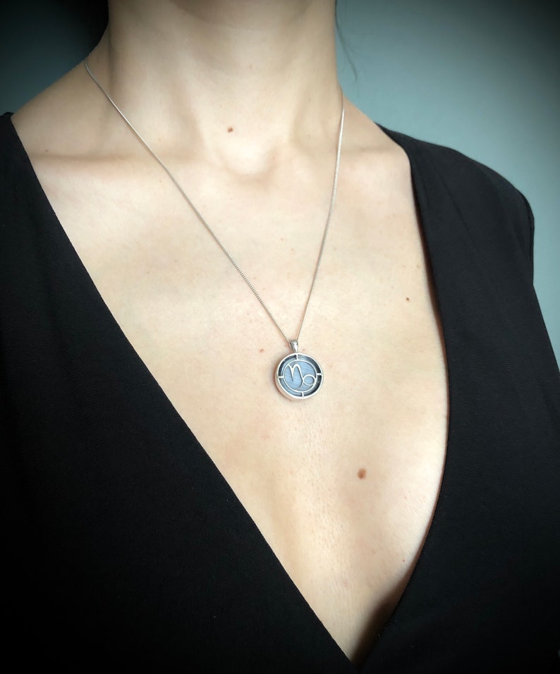 Op de afbeelding: Een zilveren ketting met een ronde hanger met het astrologische symbool voor Steenbok. De hanger is zilver met een zwarte achtergrond en het symbool is wit.