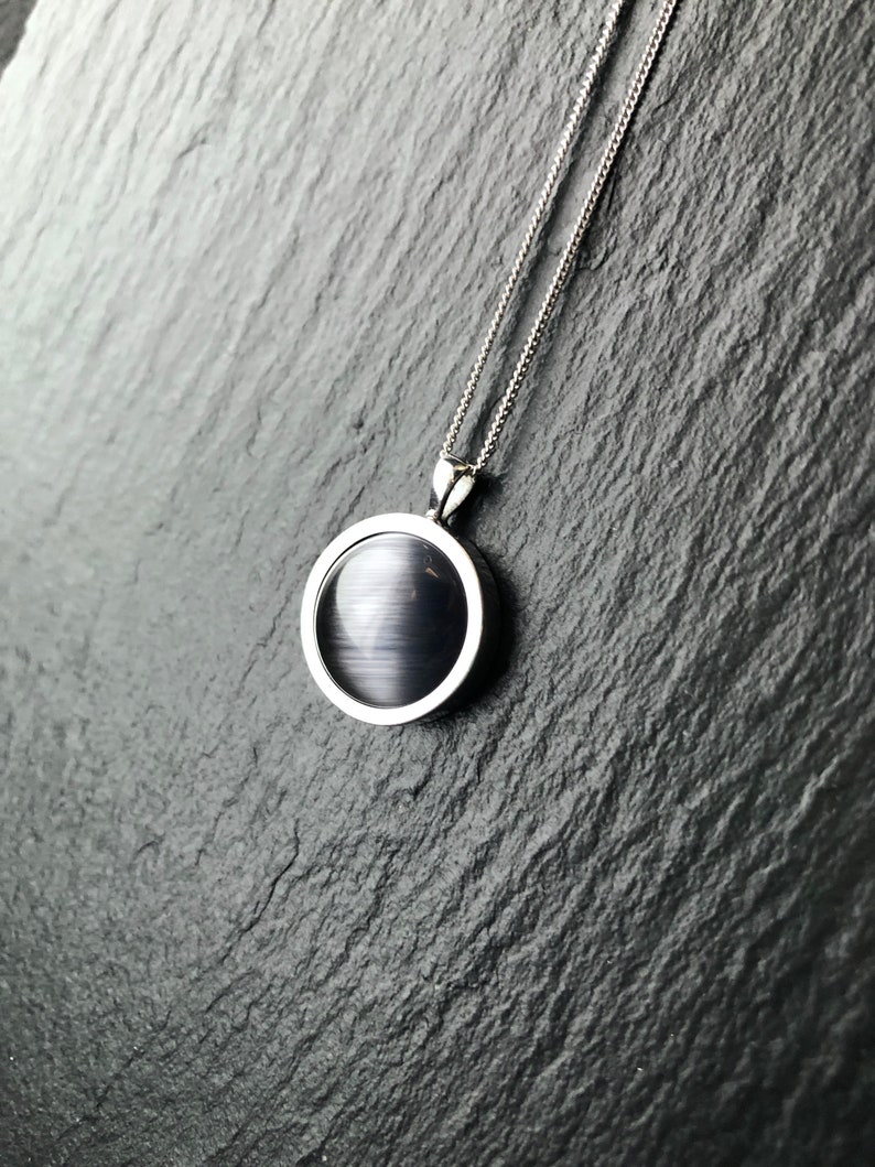 Op de afbeelding: Een zilveren ketting met een zwarte ovale hanger. De hanger heeft een glad, glanzend oppervlak.