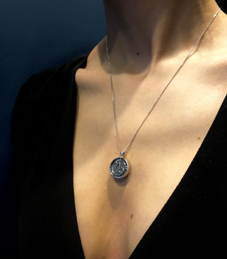 Op de afbeelding: Een zilveren ketting met een ronde hanger met het astrologische symbool voor Steenbok.