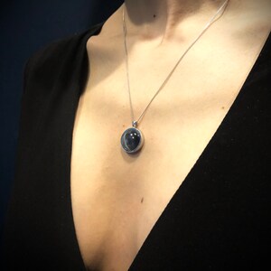 Op de afbeelding: Een zilveren ketting met een ronde hanger met een donkergrijze steen in een heldere glazen bol.